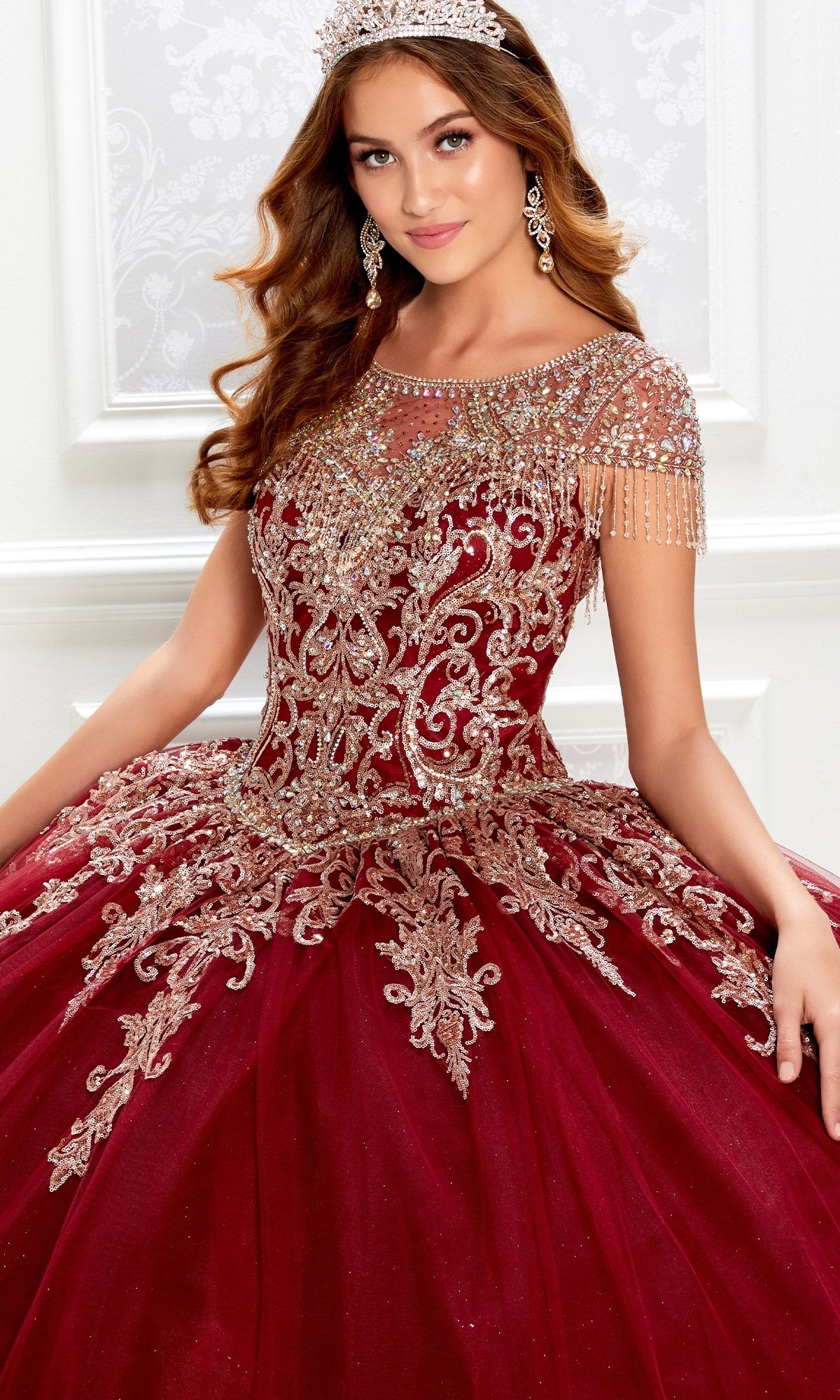 A woman wearing a jeweled tiara and earrings poses in the Princesa PR22026, a short-sleeve maroon quinceanera dress featuring a beaded bodice, intricate gold embroidery, and sparkling beadwork.